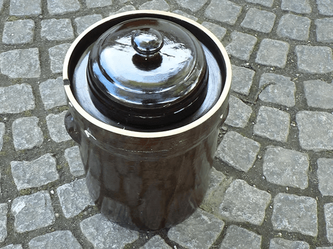 Ein brauner Steinkrug mit Deckel steht auf Kopfsteinpflaster