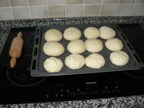 Zwölf Teigkugeln liegen auf einem Backblech, daneben ein Nudelholz